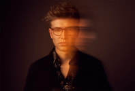 Ein Mann mit Brille, verschwommenes Doppelbild effekt, dunkler Hintergrund, wirkt nachdenklich und modern.