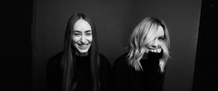 Zwei Frauen in schwarzer Kleidung lachen in die Kamera. Die linke hat langes glattes Haar, die rechte kinnlanges welliges Haar.