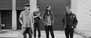 Vier Männer stehen vor einer Garagentür, alle lächeln oder lachen. Sie tragen lässige Kleidung. Schwarz-Weiß-Foto.