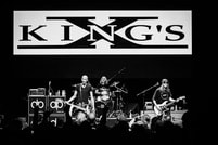 Eine Band spielt auf einer Bühne unter einem großen Schild mit der Aufschrift 'KING'S X'. Sie sind von Musikinstrumenten umgeben.