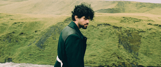 Eine Person mit lockigem Haar trägt einen grünen Mantel und steht vor einer weitläufigen, grünen Hügellandschaft.