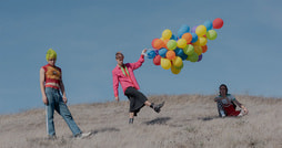 Drei Personen stehen auf einem Hügel mit trockenem Gras unter blauem Himmel. Eine Person hält bunte Luftballons, die andere trägt ein gelbes Kopftuch und eine sitzt auf dem Boden.