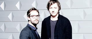 Zwei Männer in Anzügen stehen vor einer grauen Wand mit geometrischen Mustern. Der linke trägt eine Brille, der rechte hat dunkles Haar.