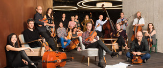 Eine Gruppe von Musiker:innen sitzt in einem modernen Raum, umgeben von Instrumenten wie Celli und Geigen, vor einem Holzhintergrund.