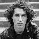 Ein schwarz-weiß Foto einer Person mit lockigem Haar und Lederjacke, die ernst in die Kamera blickt, mit Treppen im Hintergrund.