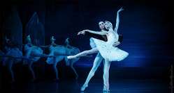 Ein Ballettpaar tanzt anmutig in weißen Kostümen auf der Bühne, flankiert von einer Gruppe von Ballerinas im Hintergrund.