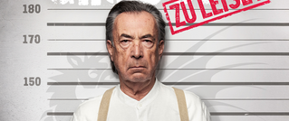 Ein älterer Mann in weißem Hemd steht vor einer Verbrecherkartei mit Markierungen in Zentimetern. Oben rechts steht 'Zu Leise' in Rot.