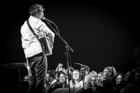 Ein Musiker spielt auf der Bühne Gitarre und singt ins Mikrofon, während ein begeistertes Publikum zuhört. Schwarz-weiß Bild.