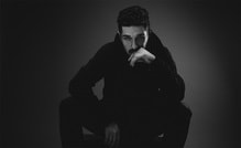 Ein Mann in einem dunklen Raum, in Schwarz-Weiß, sitzt nachdenklich mit verschränkten Armen und Hand vorm Gesicht auf einem Stuhl.