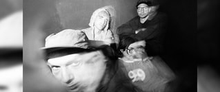 Verschwommenes Schwarz-Weiß-Bild von vier Personen. Drei sitzen, eine Person steht. Unklare Gesichtszüge und verwischte Bewegungen.