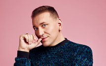 Mann mit kurzem Haar in blau gemustertem Pullover vor rosa Hintergrund. Er schaut nachdenklich, mit der Hand am Kinn.