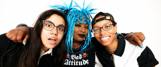 Drei Personen posieren lächelnd zusammen; die mittlere hat blaue, stachelige Haare und trägt ein 'Bad Attitude'-Shirt.