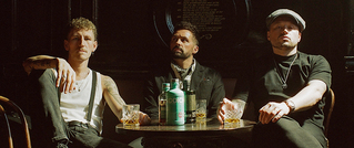 Drei Männer sitzen an einem runden Tisch in einer Bar, mit Gläsern und einer Flasche Gin darauf. Sie tragen lässige, schicke Kleidung.