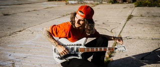 Person mit rotem T-Shirt und Mütze sitzt auf einem urbanen Boden und spielt eine weiße E-Gitarre unter Sonnenschein.