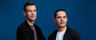 Zwei Männer in schwarzen Jacken stehen vor einem blauen Hintergrund und schauen in die Kamera.