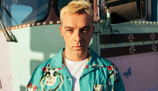 Ein Mensch mit blondem Haar trägt ein blaues Hemd mit Blumenmuster und steht vor einem pastellfarbenen Fahrzeug bei Sonnenlicht.