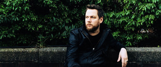 Ein Mann mit Bart sitzt in schwarzer Jacke auf einer Betonmauer vor einer grünen Hecke und schaut nach links.