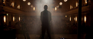 Ein silhouettierter Mann steht auf einer Bühne vor einem Publikum in einem beleuchteten Saal.