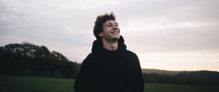 Ein lächelnder Mann steht in einer offenen Landschaft, trägt einen schwarzen Hoodie. Der Himmel ist bewölkt und die Umgebung grün.