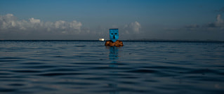 Person schwimmt in ruhigem Wasser bei Sonnenuntergang, trägt eine blaue Maske. Horizont mit Wolken ist sichtbar.