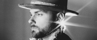 Schwarzweiß-Porträt eines bärtigen Mannes mit Hut. Ein heller Lichtstrahl scheint hinter seinem Kopf hervor.