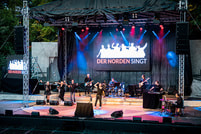 Eine beleuchtete Bühne mit Musikern und Sängern; ein großes Schild zeigt 'Der Norden Singt'.