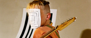 Person mit weißer Maske über dem Gesicht, hält eine elektrische Gitarre. Im Hintergrund sind schwarz-weiße Streifen zu sehen.