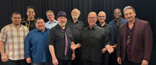 Eine Gruppe von zehn Männern posiert vor einem schwarzen Hintergrund, lächelnd und teils in bunter, teils in dunkler Kleidung.