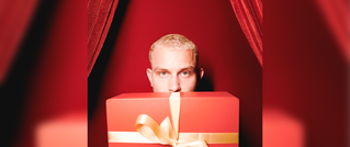 Eine Person mit kurzem blondem Haar schaut hinter einem großen roten Geschenk mit goldener Schleife hervor, vor rotem Vorhang.