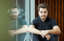 Ein Mann in einem schwarzen T-Shirt sitzt entspannt neben einer Glaswand, die sein Spiegelbild zeigt, mit Holzwand im Hintergrund.
