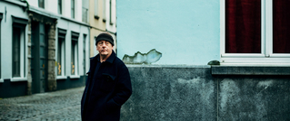 Ein älterer Mann in dunkler Kleidung steht in einer gepflasterten Gasse vor einer blauen Wand mit roten Vorhängen.