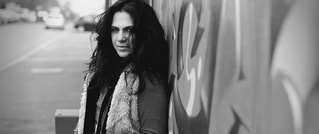 Schwarz-weiß-Foto einer Frau mit lockigem Haar, die an eine bemalte Wand gelehnt steht. Die Straße ist im Hintergrund unscharf.