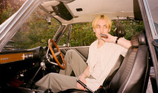 Eine Person sitzt in einem Auto, hält ein Handy ans Ohr und blickt nach draußen. Der Innenraum ist retro gestaltet.