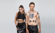 Ein Mann mit Tattoos und eine Frau mit schwarzem Top und Lederrock posieren vor neutralem Hintergrund. Beide schauen ernst.