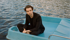 Eine Person in dunkler Kleidung sitzt in einem blauen Tretboot auf einem See, umgeben von Wasser.
