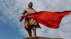 Person mit beiger Kleidung steht auf einem Felsen, hält ein rotes Tuch im Wind. Himmel voller Wolken im Hintergrund.