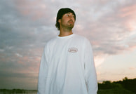 Ein Mann mit Mütze und weißem Langarmshirt steht draußen vor einem bewölkten Himmel bei Sonnenuntergang und schaut zur Seite.