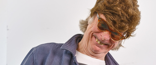 Ein lächelnder älterer Mann mit brauner Perücke und Sonnenbrille neigt seinen Kopf zur Seite, vor einem schlichten Hintergrund.
