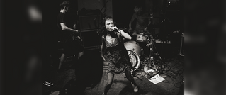 Eine Person singt leidenschaftlich in ein Mikrofon, begleitet von einer Band mit Gitarre und Schlagzeug auf einer Bühnenperformance.