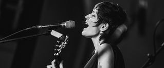 Schwarz-weiß-Foto einer singenden Person mit Kurzhaarfrisur, die Gitarre spielt und in ein Mikrofon singt.