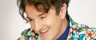 Ein lächelnder Mann mit lockigem Haar trägt ein buntes Hemd mit Blumenmuster vor einem hellen Hintergrund.