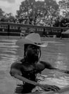 Eine Person mit Cowboyhut steht in einem Pool, umgeben von Bäumen und einem Gebäude im Hintergrund. Das Bild ist in Schwarz-Weiß.