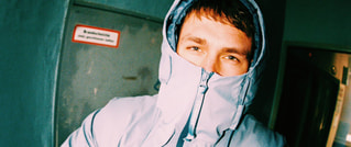 Eine Person in blauer Jacke mit Kapuze, die fast das ganze Gesicht bedeckt. Im Hintergrund ist eine graue Wand mit einem Schild.