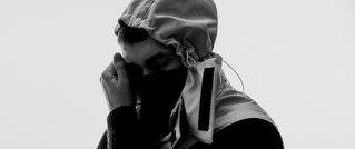 Person in Schwarz-Weiß, trägt Kapuze und Maske, hält eine Hand ans Gesicht. Hintergrund ist schlicht weiß.