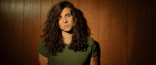 Person mit langen, lockigen Haaren in grünem T-Shirt steht vor einer Holzvertäfelung, ausdrucksvoller Blick, warmes Licht.