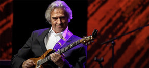 Ein Mann in einem Anzug spielt konzentriert Gitarre auf einer Bühne mit rotem Hintergrund. Ein Mikrofonständer ist sichtbar.