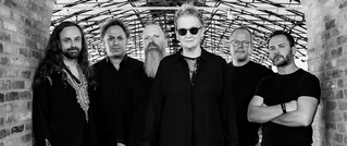 Sechs Personen stehen in einer Reihe unter einem Bogendach, alle in dunkler Kleidung. Die Szene ist in Schwarz-Weiß gehalten.