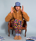 Eine Person sitzt mit erhobenen Händen in einem Sessel, trägt bunte Blumenhosen, einen braunen Hoodie und einen blauen Hut.
