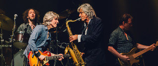 Vier Musiker auf einer Bühne: Ein Gitarrist, ein Saxophonist, ein Bassist und ein Schlagzeuger, alle in Aktion beim Auftritt.