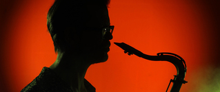 Silhouette einer Person mit Brille, die Saxofon spielt, vor einem leuchtend orange-roten Hintergrund.